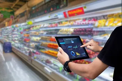 Mann steht beim Kühlregal in einem Supermarkt, hält ein Tablet in den Händen und zeigt mit seinem rechten Zeigefnger auf die Statistik des intelligenten Einzelhandelsmanagementsystems auf dem Bildschirm.
