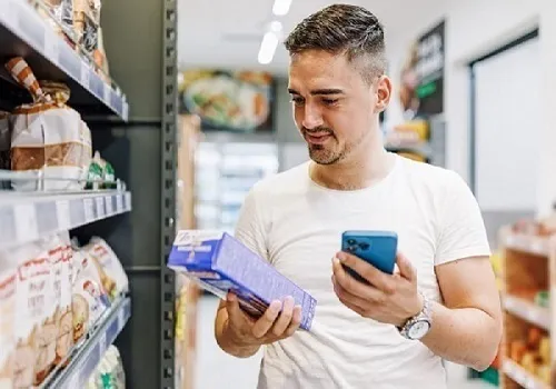 Junger Mann steht am Regal im Supermarkt, schaut ein Produkt in seiner rechten Hand an und hält in der linken Hand ein Smartphone