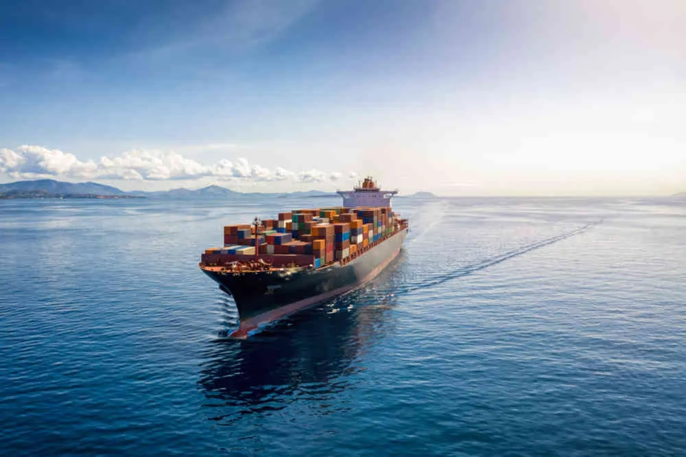 Luftaufnahme eines schwer beladenen Containerfrachtschiffs, das über ruhiges, blaues Meer fährt. R&P zeigt Ihnen hier ein Projektbeispiel.
