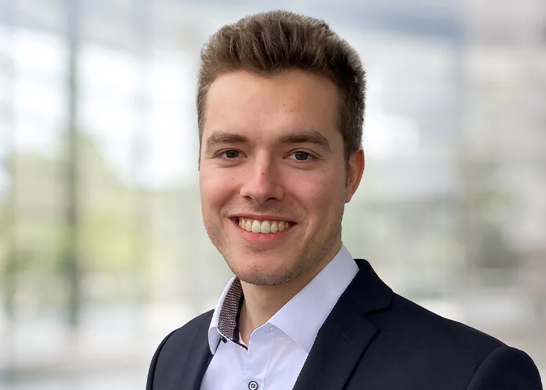 Praktikant Patrick trägt ein weißes Hemd mit einem schwarzen Jackett und lächelt breit