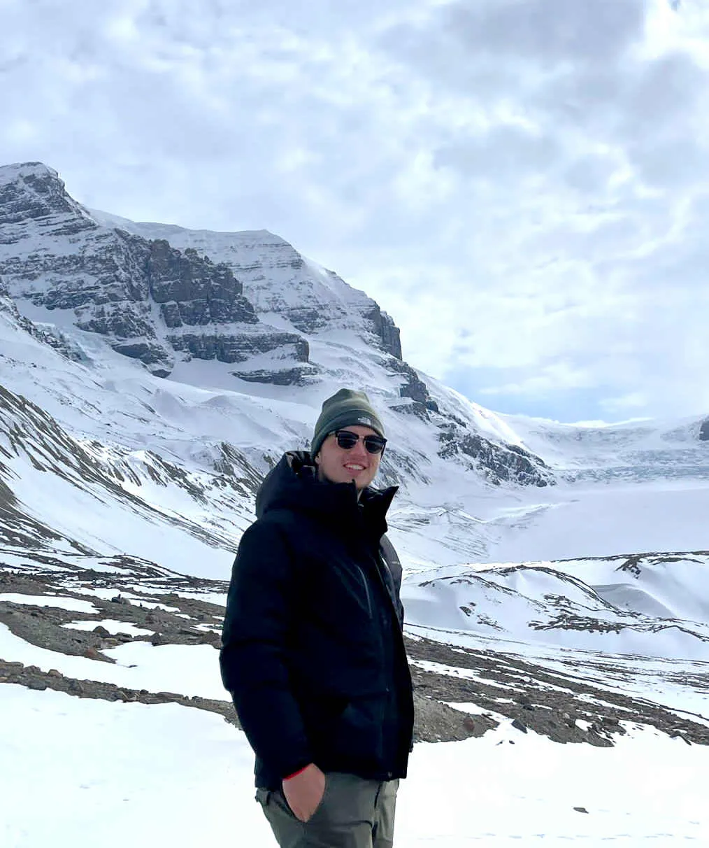 Urlaubsbild von Praktikant Sebastian zeigen ihn mit Mütze, Sonnenbrille und dicker Jacke vor schneebedeckten Bergen