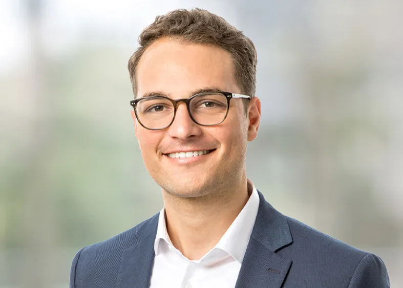 Consultant Simon trägt ein weißes Hemd mit einem dunkel blauem Jackett und lächelt breit