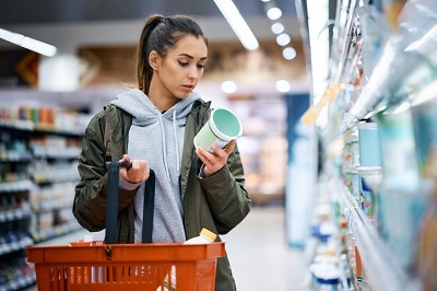 Frau steht im Supermarkt mit rotem Einkaufskorb in der Hand bei einem Regal und studiert ein Produkt in ihrer linken Hand