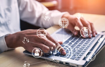 Person arbeitet am Laptop, Icons des E-Commerce werden als Hologramme über der Tastatur angezeigt