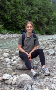 Consultant Carolin ist am R&P Standort Berlin gestartet. Sie ist gerne an der frischen Luft und zeigt sich hier beim Wandern in den Bergen, sitzend bei einem Bach.