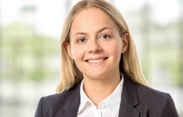 Consultant Ann-Sophie trägt eine weiße Bluse mit schwarzem Jackett und lächelt breit