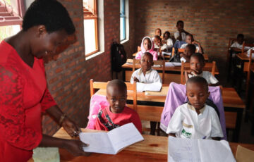 Frau in rotem Oberteil unterrichtet Grundschüler an Zweiertischen in Afrika