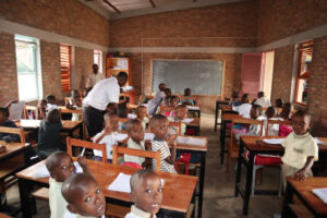 Schulklasse an Tischen in Afrika mit vielen Kindern und zwei Erwachsenen, die etwas erklären