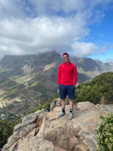 Consultant Simon steht auf einem Berg mit toller Aussicht auf Berge in roter Jacke und kurzer Hose mit Händen in den Hosentaschen