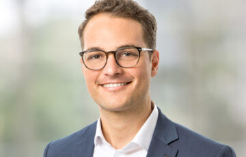 Consultant Simon trägt ein weißes Hemd mit einem blauem Jackett und lächelt breit