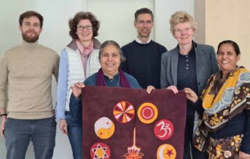 Sechs lächelnde Personen halten Flagge mit Symbolen verschiedener Religionen hoch: Besuch von MAHER und Indienhilfe e.V. (Kelkheim) im Kölner R&P Office