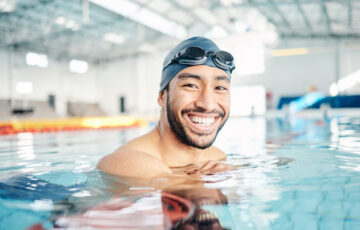 Mann mit Schwimmbrille schaut im Hallenbad lächelnd in die Kamera