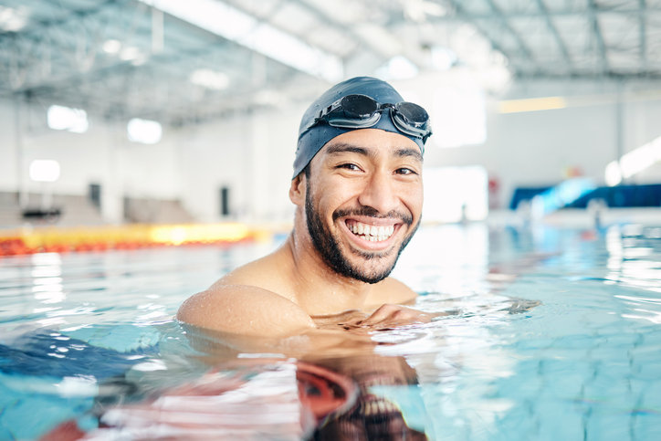 Mann mit Schwimmbrille schaut im Hallenbad lächelnd in die Kamera