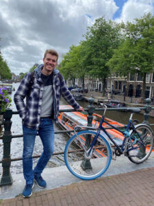 Praktikant Patrick steht auf einer Brücke neben einem Fahrrad, während ein rotes Boot durch die Brücke fährt