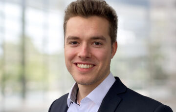 Praktikant Patrick trägt ein weißes Hemd mit einem schwarzen Jackett und lächelt breit